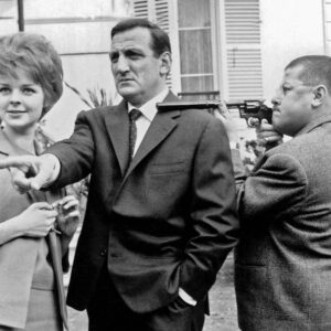 Photographie de plateau en noir et blanc des Tontons Flingueurs : Lino Ventura pointe du doigt aux côtés de Sabine Sinjen, tandis que Francis Blanche vise avec un revolver à silencieux devant une maison.