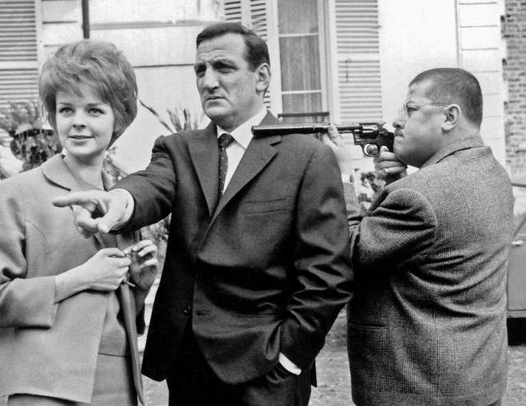 BRLV108 - 1648319 Photographie de plateau en noir et blanc des Tontons Flingueurs : Lino Ventura pointe du doigt aux côtés de Sabine Sinjen, tandis que Francis Blanche vise avec un revolver à silencieux devant une maison.