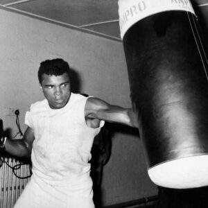 Affiche Mohamed Ali : photographie d'action en noir et blanc de 1963 montrant Mohamed Ali (Cassius Clay) portant un coup puissant dans un sac de frappe historique "Ampro London" en cuir.