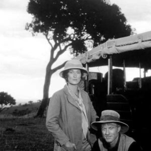 Photographie de plateau en noir et blanc du film Out of Africa montrant Meryl Streep et Robert Redford en tenue de safari. Meryl Streep est debout, les mains dans les poches de sa veste, tandis que Robert Redford est accroupi à ses côtés. Ils posent devant un véhicule de brousse et un grand acacia dans un paysage de plaine africaine.