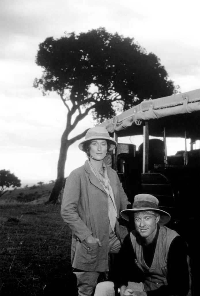 BRMSTR100NB Photographie de plateau en noir et blanc du film Out of Africa montrant Meryl Streep et Robert Redford en tenue de safari. Meryl Streep est debout, les mains dans les poches de sa veste, tandis que Robert Redford est accroupi à ses côtés. Ils posent devant un véhicule de brousse et un grand acacia dans un paysage de plaine africaine.