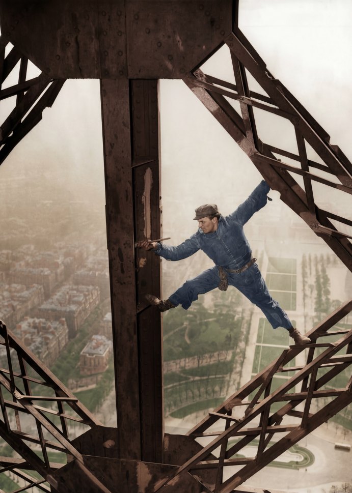 L'Acrobate de 1953 : Le Peintre de la Tour Eiffel | Fuzars Photographie historique colorisée de 1953 montrant un peintre en bâtiment en équilibre sur la structure de la Tour Eiffel, un pinceau à la main, surplombant Paris.