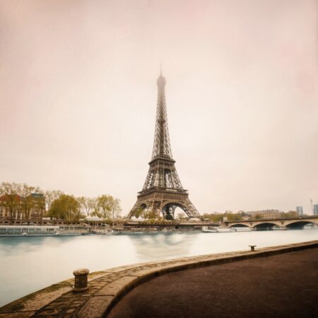 Photographie artistique colorisée de la Tour Eiffel vue depuis les quais de Seine. L'eau présente un aspect vaporeux (pose longue) et le ciel est baigné d'une lumière douce et brumeuse.