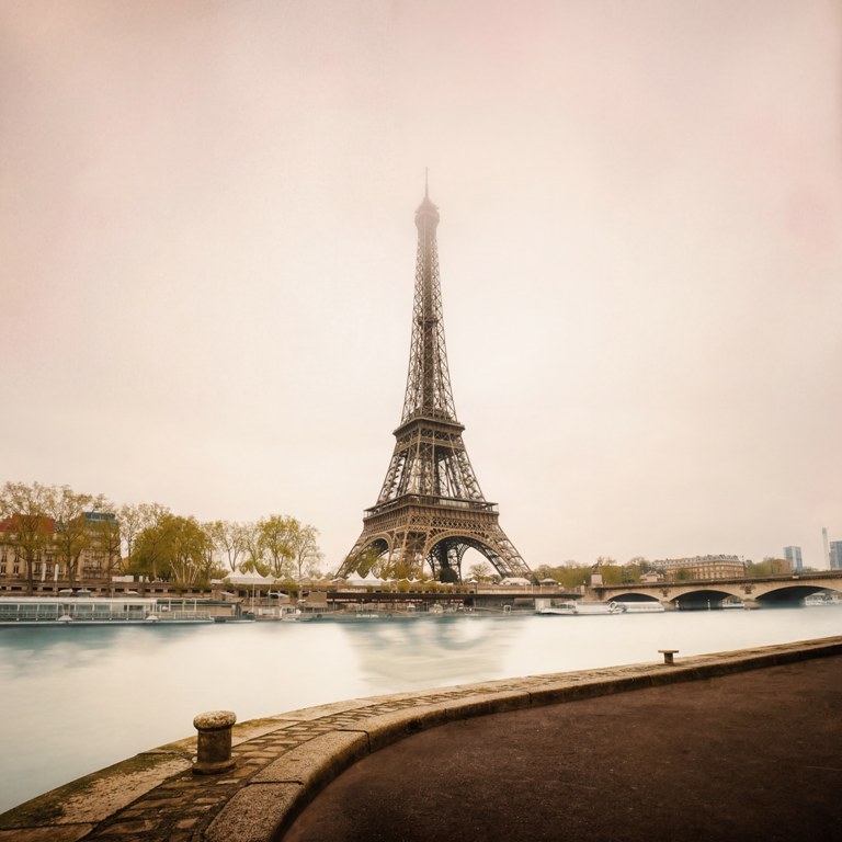 Photographie artistique colorisée de la Tour Eiffel vue depuis les quais de Seine. L'eau présente un aspect vaporeux (pose longue) et le ciel est baigné d'une lumière douce et brumeuse.