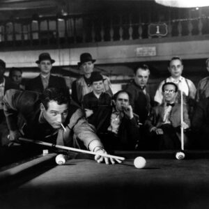 Photographie de plateau en noir et blanc du film L'Arnaqueur (1961) montrant Paul Newman penché sur une table de billard, une cigarette aux lèvres, s'apprêtant à tirer sous le regard d'un groupe d'hommes dans une salle de jeu sombre.