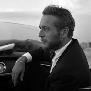 Photographie de profil en noir et blanc de Paul Newman en 1963 à Venise. L'acteur, portant une barbe courte et vêtu d'un costume élégant avec nœud papillon, regarde vers l'horizon depuis un bateau naviguant sur la lagune.