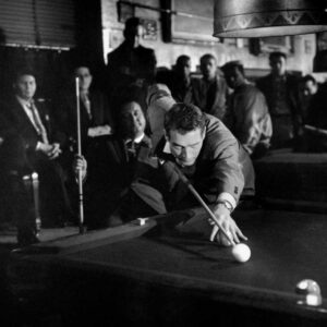 Photo noir et blanc du film L'Arnaqueur (1961) montrant Paul Newman en pleine action, le corps très incliné au-dessus d'une table de billard pour préparer un coup de queue. Un groupe d'hommes l'observe avec attention dans l'ombre d'une salle de jeu enfumée.