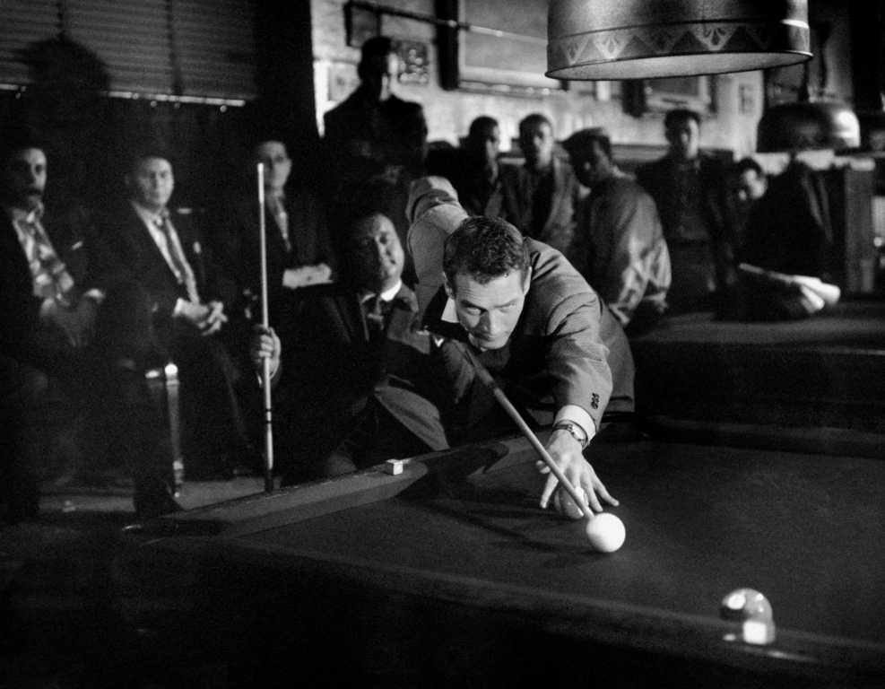 BRPN105NB 2628745 Photo noir et blanc du film L'Arnaqueur (1961) montrant Paul Newman en pleine action, le corps très incliné au-dessus d'une table de billard pour préparer un coup de queue. Un groupe d'hommes l'observe avec attention dans l'ombre d'une salle de jeu enfumée.