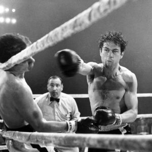 Photographie en noir et blanc du film Raging Bull montrant Robert De Niro (Jake LaMotta) sur un ring de boxe, en plein effort, assénant un coup de poing puissant à son adversaire sous le regard attentif de l'arbitre. La scène est marquée par un éclairage dramatique et des projections de sueur capturées en plein mouvement.