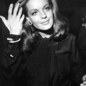 Photographie en noir et blanc de l'actrice Romy Schneider souriante lors d'un événement mondain, la main délicatement relevée près de son visage, portant un élégant chemisier sombre noué au col et des bijoux scintillants.