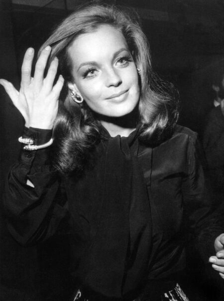 Photographie en noir et blanc de l'actrice Romy Schneider souriante lors d'un événement mondain, la main délicatement relevée près de son visage, portant un élégant chemisier sombre noué au col et des bijoux scintillants.