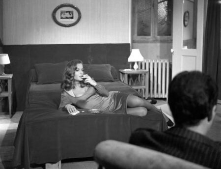 Photographie en noir et blanc de Romy Schneider dans le film Max et les Ferrailleurs, allongée sur un lit, vêtue d'une robe et fumant une cigarette, le regard tourné vers un homme assis de dos au premier plan dans une chambre.