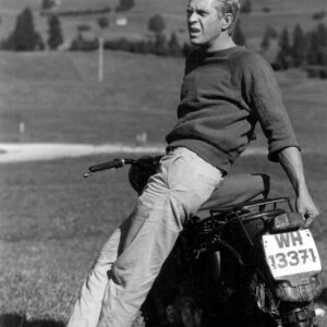 Photographie en noir et blanc de Steve McQueen dans le film La Grande Évasion. L'acteur est nonchalamment appuyé contre une moto, portant son célèbre sweatshirt à manches courtes et un pantalon clair, regardant vers l'horizon dans un paysage de campagne vallonné.