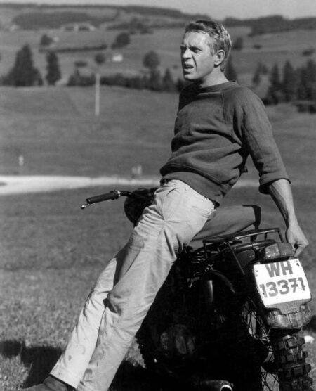 Photographie en noir et blanc de Steve McQueen dans le film La Grande Évasion. L'acteur est nonchalamment appuyé contre une moto, portant son célèbre sweatshirt à manches courtes et un pantalon clair, regardant vers l'horizon dans un paysage de campagne vallonné.