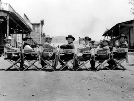 Photo panoramique noir et blanc du casting du film Les Sept Mercenaires assis sur des chaises de réalisateur : Steve McQueen, Yul Brynner, Charles Bronson et les autres acteurs en tenues de cowboys.