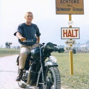 Photographie en couleur de l'acteur Steve McQueen vêtu d'un pull bleu, assis sur une moto vintage devant des panneaux jaunes de la frontière suisse, tirée du film La Grande Évasion.