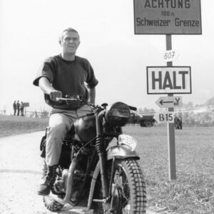 1638434 THE GREAT ESCAPE, 1963 directed by JOHN STURGES Steve Mc Photographie en noir et blanc de l'acteur Steve McQueen assis sur une moto vintage, arrêté devant des panneaux en allemand indiquant la frontière suisse, tirée du film La Grande Évasion.
