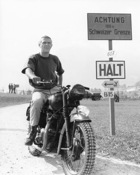 Photographie en noir et blanc de l'acteur Steve McQueen assis sur une moto vintage, arrêté devant des panneaux en allemand indiquant la frontière suisse, tirée du film La Grande Évasion.