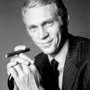 Portrait en noir et blanc de Steve McQueen dans le film L'Affaire Thomas Crown. L'acteur, très élégant en costume à rayures et cravate, regarde l'objectif avec un sourire en coin tout en tenant un cigare entre ses mains jointes.