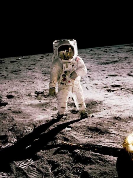 Photographie historique haute définition d'un astronaute de la NASA debout sur la surface de la Lune, tenant un instrument scientifique. Le module lunaire LEM et le drapeau américain sont reflétés dans sa visière dorée.
