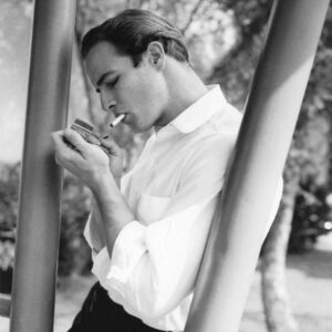 Portrait de profil en noir et blanc de Marlon Brando jeune allumant une cigarette, vêtu d'une chemise blanche et appuyé contre un poteau en extérieur.