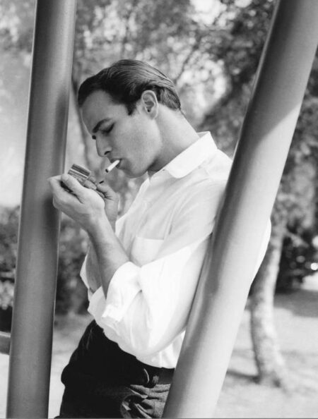 Portrait de profil en noir et blanc de Marlon Brando jeune allumant une cigarette, vêtu d'une chemise blanche et appuyé contre un poteau en extérieur.
