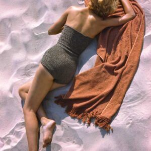 Photographie vintage colorisée d'un mannequin femme en tenue élégante des années 1940 (grand chapeau de paille, jupe rayée) debout sur une vaste plage de sable face à l'océan.
