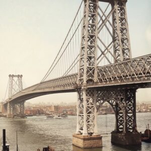 Photographie historique colorisée de 1903 montrant le Pont de Williamsburg à New York, l'année de son ouverture. La structure massive en acier s'étend sur l'East River, reliant Manhattan et Brooklyn. Des calèches à cheval, des tramways électriques et une foule de piétons traversent le pont sous un ciel dégagé. Le fleuve est animé par des bateaux à vapeur et des barges.