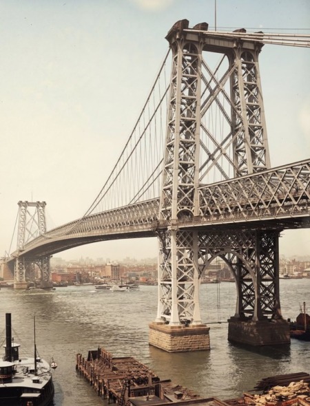 Photographie historique colorisée de 1903 montrant le Pont de Williamsburg à New York, l'année de son ouverture. La structure massive en acier s'étend sur l'East River, reliant Manhattan et Brooklyn. Des calèches à cheval, des tramways électriques et une foule de piétons traversent le pont sous un ciel dégagé. Le fleuve est animé par des bateaux à vapeur et des barges.