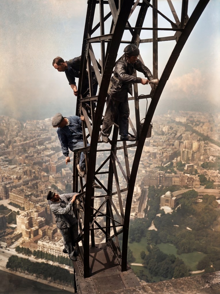 Les Équilibristes de la Tour Eiffel (1930) Photographie historique colorisée des ouvriers peintres travaillant sur la structure en fer de la Tour Eiffel, surplombant le Champ-de-Mars et Paris.