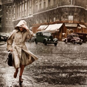 Paris 1952 : Éclats de Pluie Photographie historique colorisée d'une femme courant sous une pluie battante à Paris en 1952. Elle porte un trench-coat et une capuche. On aperçoit des voitures vintage et le Bar Lutterbach en arrière-plan.