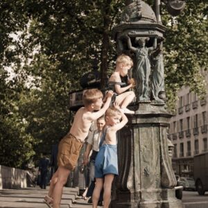 Les Garnements de la Fontaine Wallace | Fuzars Photographie d'époque colorisée montrant trois petits garçons jouant et se rafraîchissant autour d'une fontaine Wallace dans une rue de Paris. Scène de vie nostalgique.