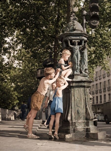 Photographie d'époque colorisée montrant trois petits garçons jouant et se rafraîchissant autour d'une fontaine Wallace dans une rue de Paris. Scène de vie nostalgique.