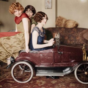 Photographie colorisée des Années Folles montrant trois jeunes femmes élégantes en robes d'époque posant avec une voiture à pédales miniature dans un salon.