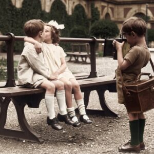 Photographie vintage colorisée de 1930 montrant un jeune garçon photographiant deux autres enfants en train de s'embrasser sur un banc dans un parc.