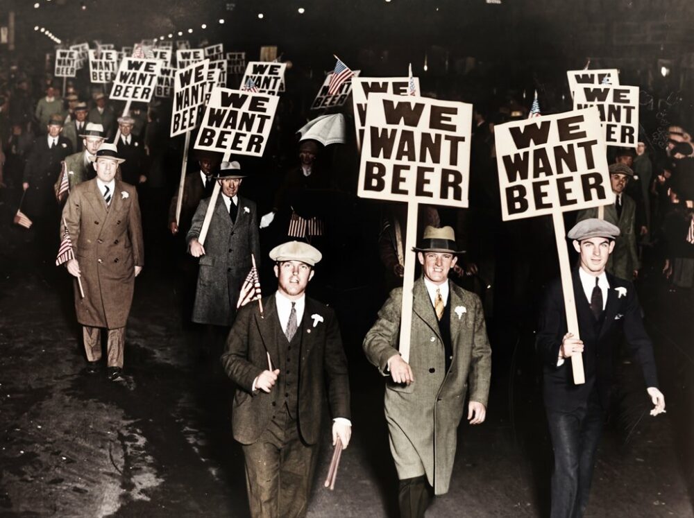 Manifestation We Want Beer : Affiche Vintage & Humour | Fuzars Photographie historique colorisée montrant une foule d'hommes en costume manifestant la nuit contre la Prohibition aux États-Unis, brandissant des panneaux "WE WANT BEER".
