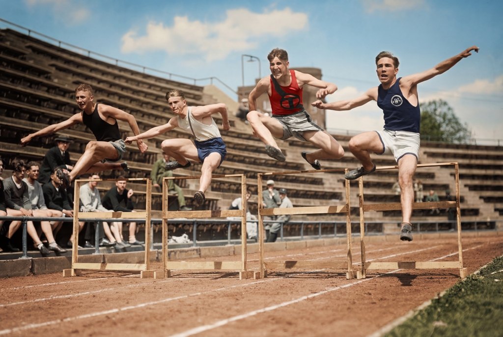 Sprint de Haies (1930) : Affiche Sport Vintage | Fuzars Photographie historique colorisée montrant quatre jeunes athlètes franchissant des haies en bois sur une piste d'athlétisme dans un stade des années 30.