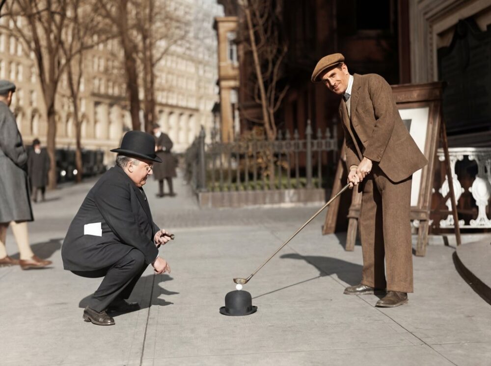 Golf de Rue (1910) : Affiche Vintage & Humour | Fuzars Photographie historique colorisée de 1910 montrant deux hommes en costume dans une rue. L'un utilise un chapeau melon pour poser une balle de golf tandis que l'autre se prépare à frapper avec son club.