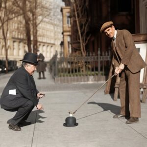 Golf de Rue (1910) : Affiche Vintage & Humour | Fuzars Photographie historique colorisée de 1910 montrant deux hommes en costume dans une rue. L'un utilise un chapeau melon pour poser une balle de golf tandis que l'autre se prépare à frapper avec son club.