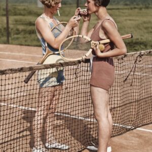 Pause au Filet : Tennis Vintage & Élégance | Fuzars Photographie historique colorisée montrant deux jeunes femmes en tenue de sport des années 1920 s'accordant une pause au filet d'un court de tennis pour allumer une cigarette, leurs raquettes en bois sous le bras.