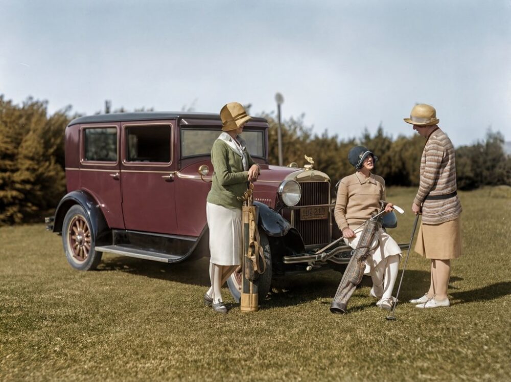 Élégance au Golf (1928) : Affiche Lifestyle Vintage | Fuzars Photographie historique colorisée des années 1920 montrant trois femmes élégantes en tenue de golf discutant près d'une voiture d'époque bordeaux dans un paysage de campagne.