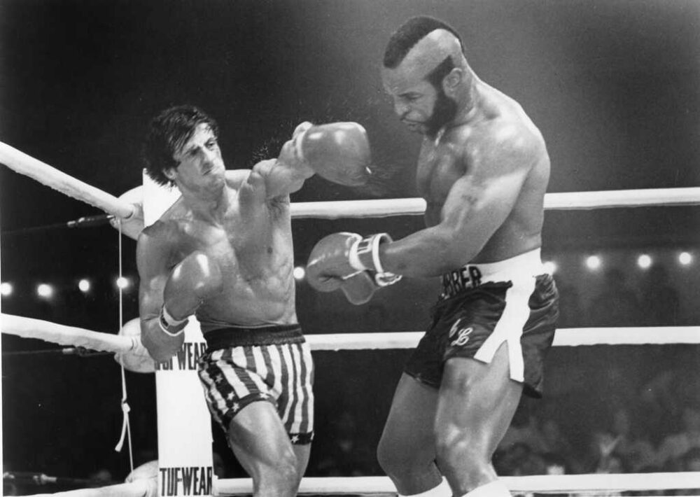 Photographie en noir et blanc de Sylvester Stallone dans son rôle emblématique de Rocky Balboa pour le film Rocky III. L'acteur est représenté torse nu, en pleine forme physique, portant ses gants de boxe et prêt au combat sur le ring.