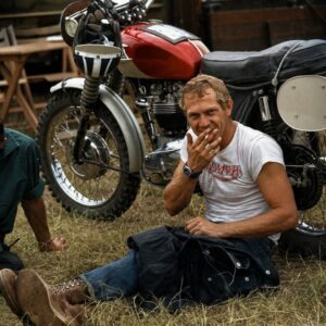 Photographie noir et blanc de l'acteur Steve McQueen souriant avec une cigarette, assis dans l'herbe devant une moto Triumph.