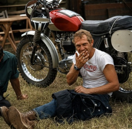 Photographie noir et blanc de l'acteur Steve McQueen souriant avec une cigarette, assis dans l'herbe devant une moto Triumph.