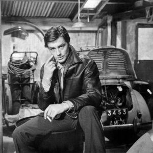 BRADEL100 -1780805 Photographie en noir et blanc de l'acteur Alain Delon portant un blouson de cuir, assis avec une cigarette à la main dans un garage automobile devant une voiture d'époque, tirée du film Mélodie en sous-sol.