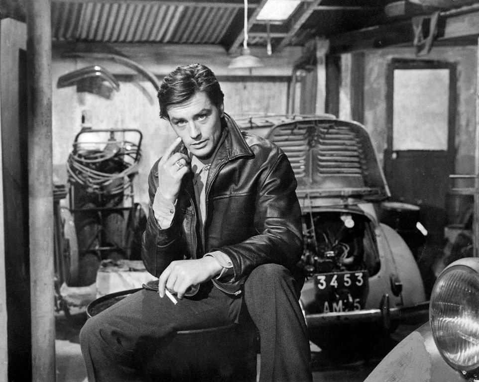 Photographie en noir et blanc de l'acteur Alain Delon portant un blouson de cuir, assis avec une cigarette à la main dans un garage automobile devant une voiture d'époque, tirée du film Mélodie en sous-sol.