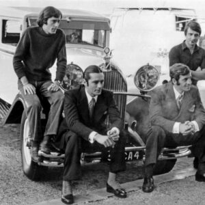 Photographie en noir et blanc d'Alain Delon et Jean-Paul Belmondo assis sur le pare-chocs d'une voiture d'époque, accompagnés de leurs frères debout derrière eux, sur les coulisses du tournage du film Borsalino.