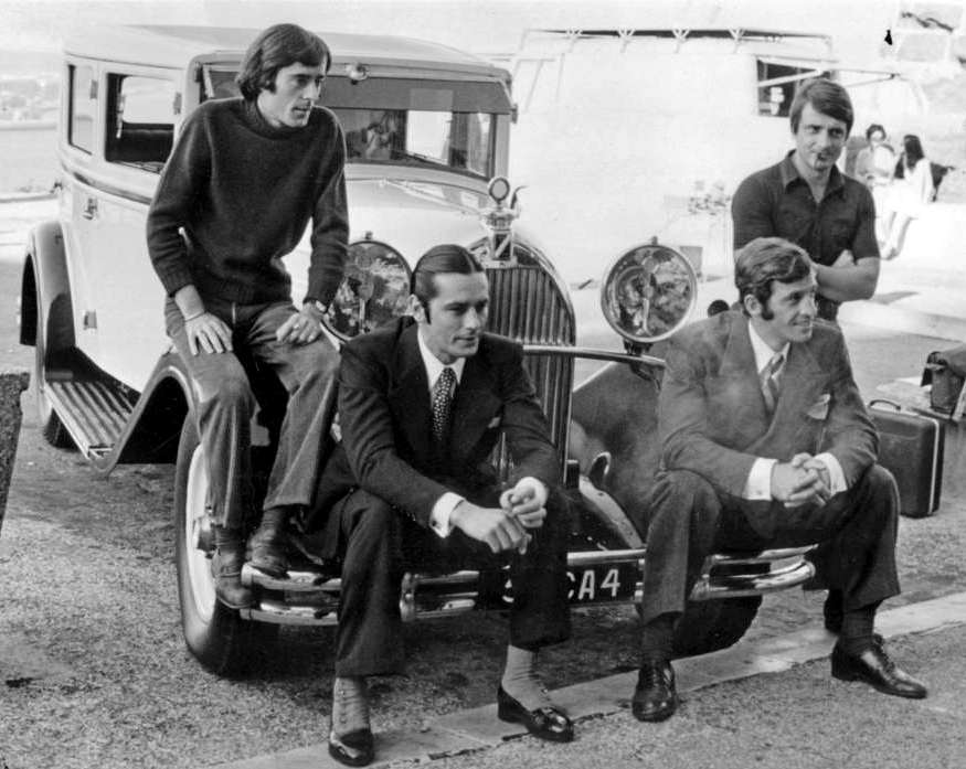 BRJPB107 - 2690787 Photographie en noir et blanc d'Alain Delon et Jean-Paul Belmondo assis sur le pare-chocs d'une voiture d'époque, accompagnés de leurs frères debout derrière eux, sur les coulisses du tournage du film Borsalino.