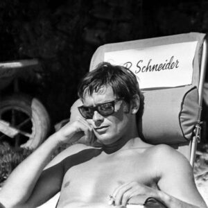 Affiche Delon La Piscine : photographie d'art en noir et blanc d'Alain Delon torse nu avec des lunettes de soleil, assis sur une chaise de tournage au nom de Romy Schneider en 1968.