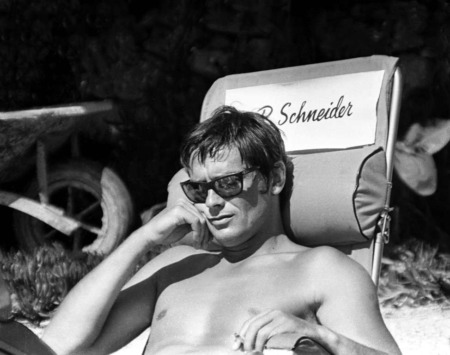 Affiche Delon La Piscine : photographie d'art en noir et blanc d'Alain Delon torse nu avec des lunettes de soleil, assis sur une chaise de tournage au nom de Romy Schneider en 1968.